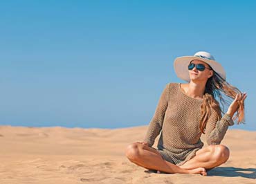 Una giovane donna seduta in spiaggia a prendere il sole con cappello e occhiali da sole per proteggersi